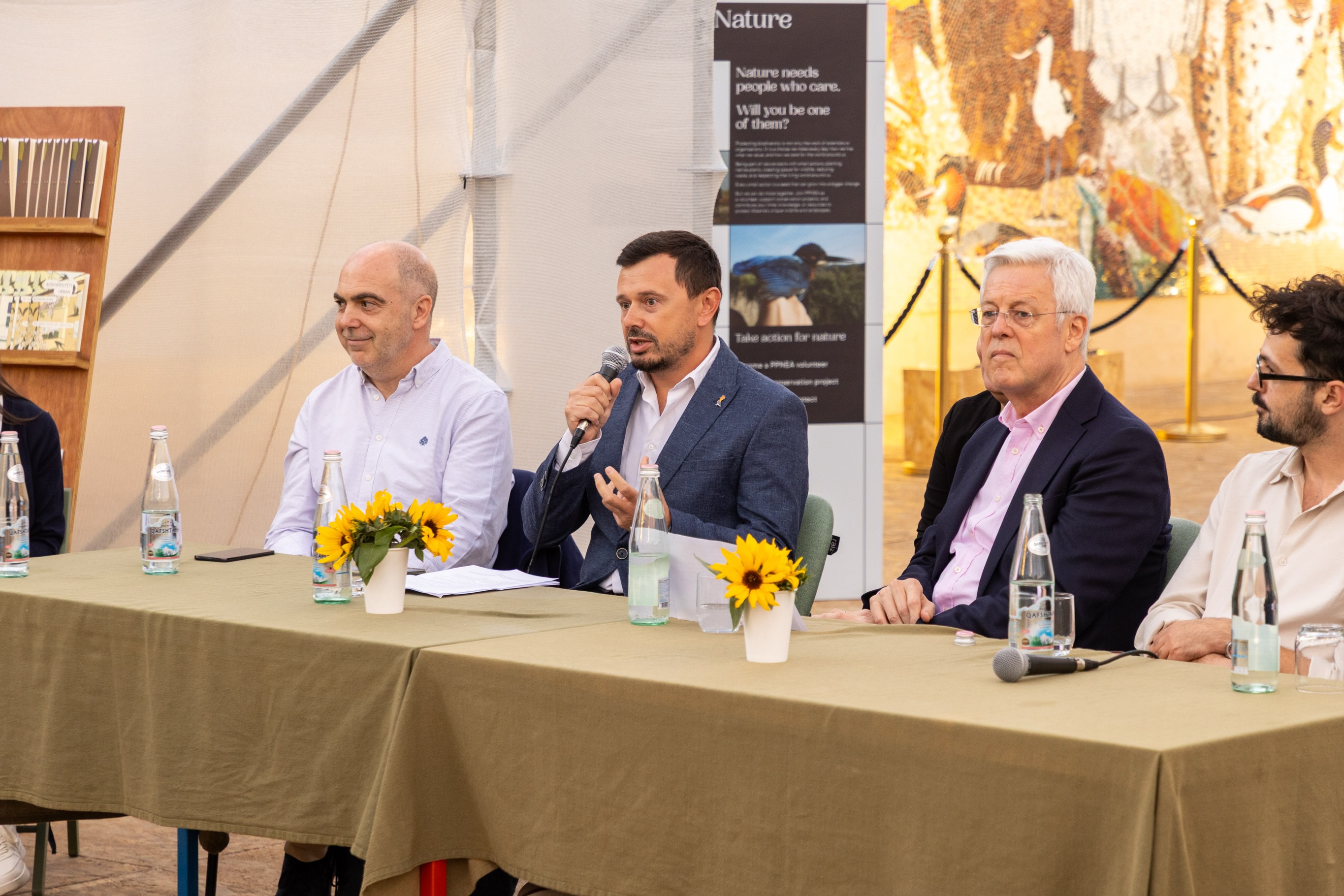 Katër folës të ulur në tryezën e panelit; njëri flet në mikrofon.